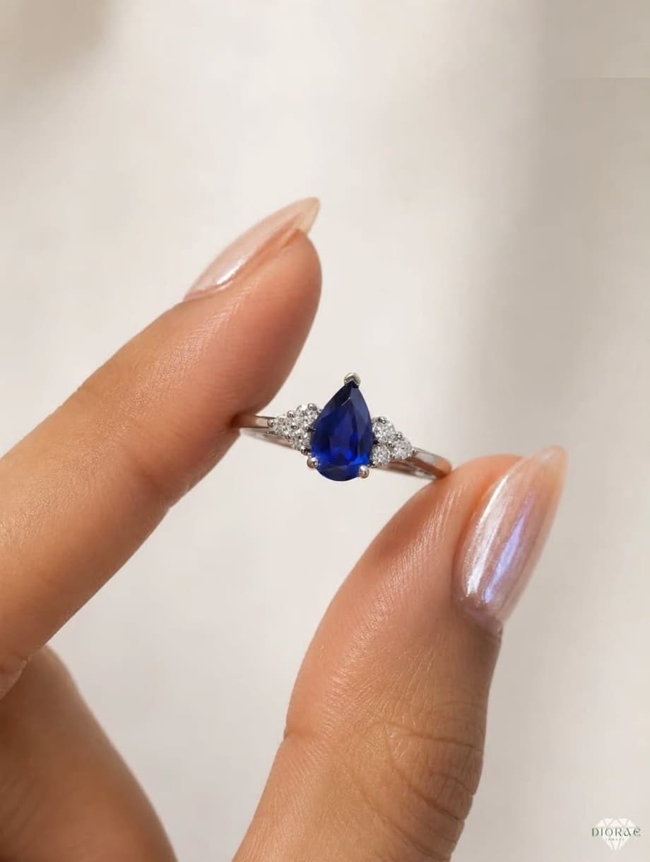 Noor sapphire ring held between two fingers, pear-cut royal blue sapphire with diamond accents on a white gold band